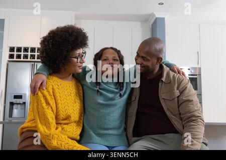 Vielfältige Familie, die auf der Küchenzeile am Edelstahl-Kühlschrank sitzt und das Kind in einem blauen Pullover umarmt Stockfoto