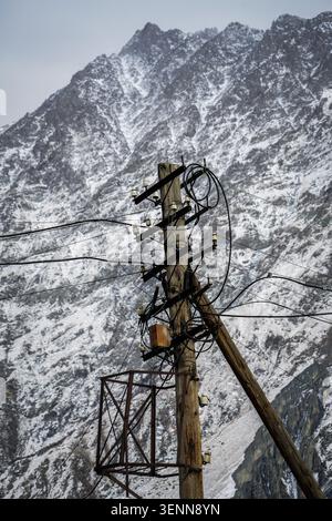 Ein rustikaler hölzerner Versorgungsmast mit einer Vielzahl von verwickelten elektrischen Drähten und weißen Keramikisolatoren steht vor dem Hintergrund eines Schnees Stockfoto