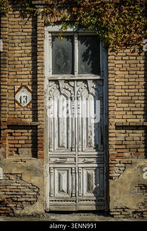 Aus nächster Nähe sehen Sie eine veraltete, verzierte Holztür mit abblätternder weißer Farbe und ein Fenster darüber, das an einer rustikalen Ziegelwand angebracht ist. Grüner und herbstlicher Efeu VI Stockfoto