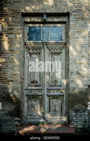Ein vertikaler Vollrahmen zeigt eine alte, verzierte Holztür mit abblätternder grauer Farbe und dekorativen Schnitzereien. Die Tür verfügt über zwei kleine Fensterscheiben Stockfoto