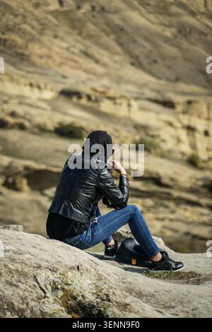 Eine Frau mit schwarzem Hijab, Lederjacke und blauer Jeans sitzt auf einem großen Felsen und blickt über die gewellten, erodierten Felsformationen der Uplists Stockfoto