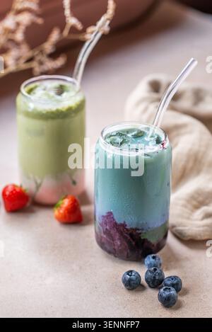 Zwei bunte Matcha-Eisgetränke, eines mit Erdbeere und eines mit Heidelbeere, serviert mit frischem Obst und Glashalmen. Stockfoto