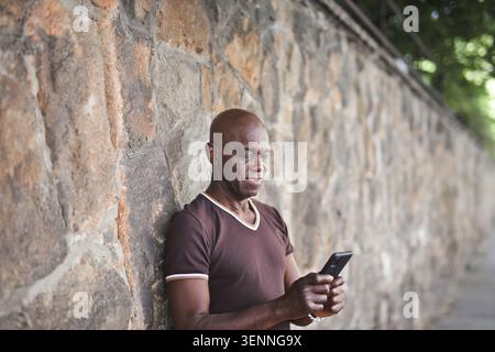 Reifer schwarzer Mann, der ein Smartphone gegen eine Steinmauer benutzt Stockfoto