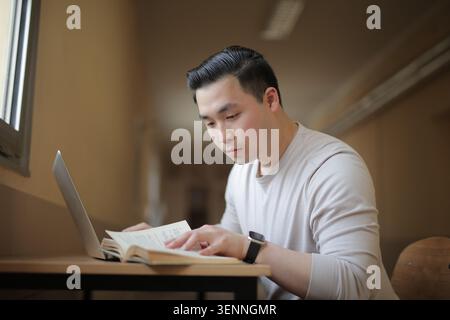 Ein fokussierter asiatischer Mann liest ein Buch an einem Schreibtisch Stockfoto