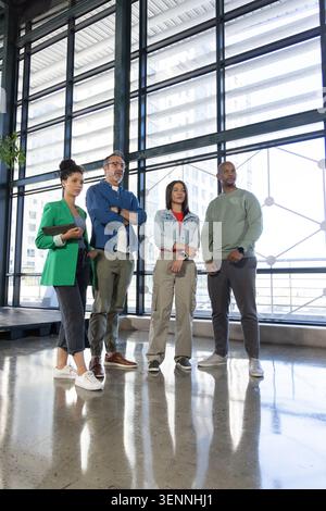 Verschiedene Kollegen posieren im modernen Atrium an Stahlfenstern in Geschäftskleidung mit Tablettenhalter Stockfoto