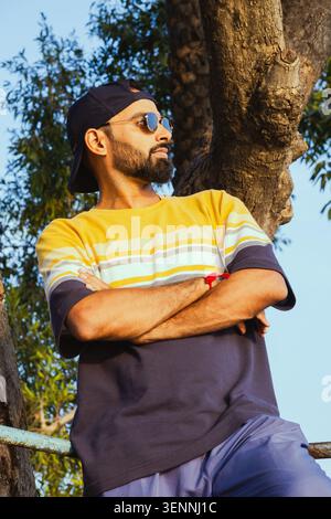 Ein junger Mann mit Bart und Sonnenbrille steht draußen mit überkreuzten Armen und blickt weg. Stockfoto