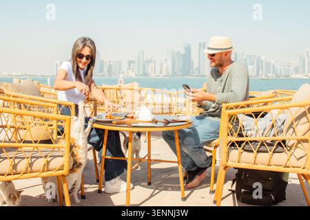 Nahaufnahme Mann und Frau sitzen im Freiluftcafé Doha Corniche. Frau spielt Katzen füttern, Mann mit Smartphone, der Spaß hat. Korbstühle, Tisch, Essen, Stockfoto