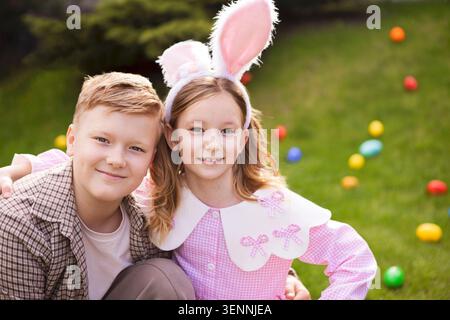 Zwei Kinder, ein Junge und ein Mädchen, posieren gemeinsam im Freien in Hasenohren und festlichen Outfits, mit bunten Ostereiern auf dem Gras Stockfoto