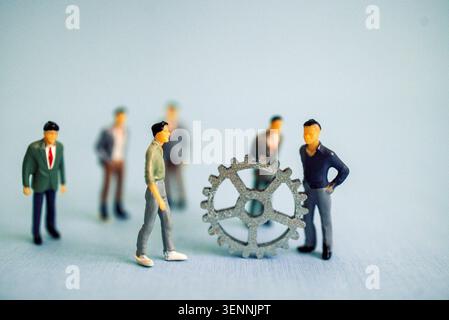 Männliche Miniaturfiguren in Anzügen und Freizeitkleidung versammeln sich um eine große Ausrüstung auf einer hellblauen Oberfläche und veranschaulichen die Zusammenarbeit in Technik und Unternehmen Stockfoto