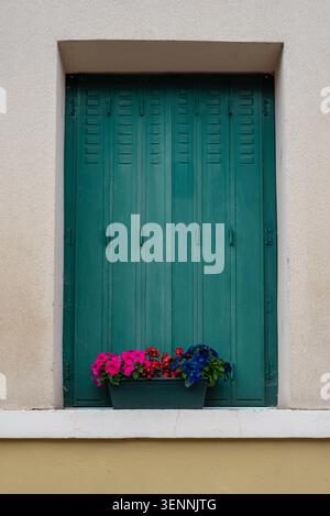 Fenster mit geschlossenen grünen Holzläden und einem Blumentopf mit blühenden Blumen auf der Fensterbank. Europäischer Dorfcharme Stockfoto