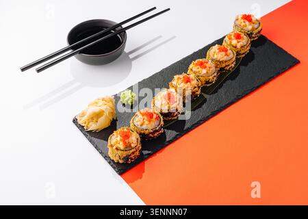 Sushi-Brötchen sind auf einem schwarzen Teller angeordnet. Jede Rolle hat rote Beläge. Es gibt eingelegten Ingwer und Wasabi an der Seite. Stäbchen sitzen neben dem Teller. Stockfoto