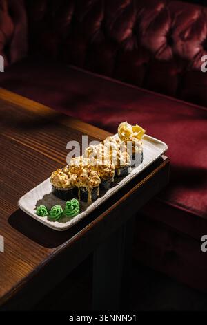 Gebackene Sushi-Brötchen auf Holztisch im Restaurant. Wasabi und eingelegter Ingwer sitzen neben den Brötchen. Die Einrichtung zeigt einen dunklen Holztisch und kuschelrote Sitzplätze im Hintergrund. Stockfoto