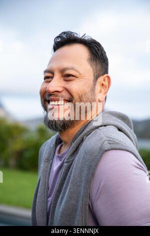 Asiatischer Mann lächelt, blickt im Freien vom Wasser weg, trägt einen grauen Hoodie und ein lila Hemd. Reif, Natur, See, Gras, bewölkt, diffus, ruhig Stockfoto