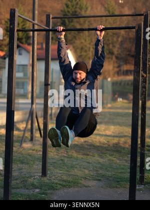 Mann, der bei kaltem Wetter an der Metallbar im Park ein Pull-up-Bein-Raise-Workout im Freien macht Stockfoto