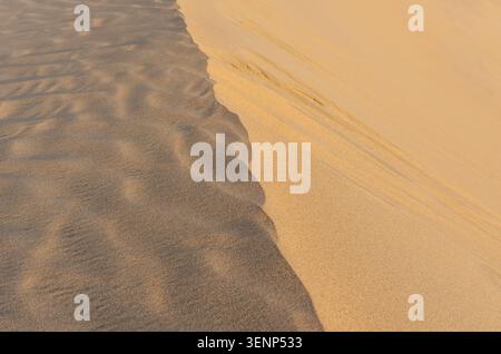 Ein abstraktes Hochwinkelbild, das den scharfen Kontrast von hellem Sonnenlicht und tiefem Schatten auf strukturierten Wüstensanddünen in Katar zeigt. Stockfoto