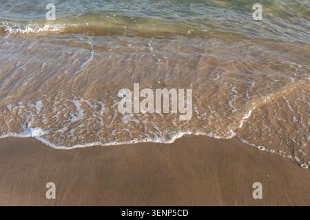 Ein Hochwinkelblick auf sanfte Meereswellen, die über einem Sandstrand schwimmen, und detaillierte weiße Meeresschaummuster an der Binnensee, Katar, erzeugen. Stockfoto