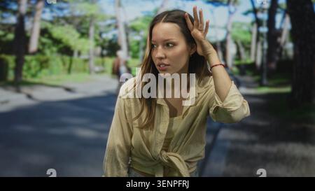 Junge Frau, die Hand an Ohr auf der Straße neben dem grünen Parkweg hält, Hemd gebunden und schielender Ausdruck; Verwirrung. Stockfoto