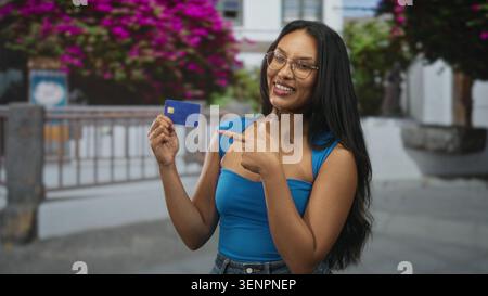 Junge Brünette, die eine Kreditkarte hält und auf der Straße mit dem Finger auf die Karte zeigt, die neben Geländer und Bougainvillea blau oben trägt; Selbstvertrauen Stockfoto