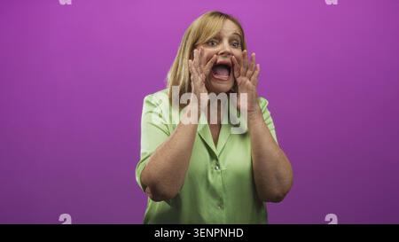 Frau mittleren Alters tönt Hand zu Mund und Gesten mit der anderen Hand im Studio in grüner Bluse vor violettem Hintergrund; dringende Kommunikation. Stockfoto