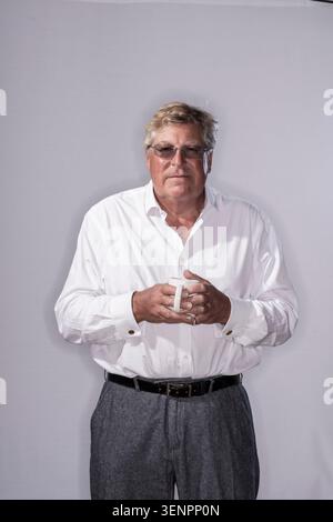 Edward Stourton steht vor einem schlichten Hintergrund und hält einen Becher. Er schaut direkt in die Kamera, während er ein weißes Hemd und eine graue Hose trägt, Creat Stockfoto