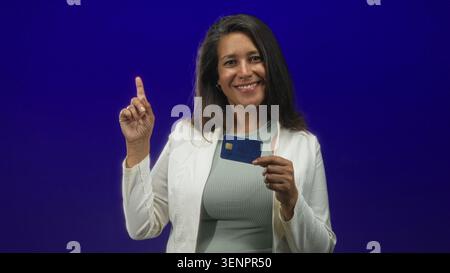 Frau, die Kreditkarte und Zeigefinger hält, während sie im Studio lächelt; Vertrauen in finanzielle Stärkung. Stockfoto