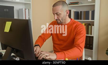 Mann in orangefarbenem Sweatshirt, der am Computer schreibt, Hände auf der Tastatur am Schreibtisch im Gebäude; Konzentration auf Produktivität. Stockfoto