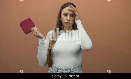 Frau, die mit der rechten Hand ein rosafarbenes Portemonnaie hält, während sie im Studio mit der linken Hand die Stirn drückt, mit weißem Oberteil und Jeans; finanzielle Frustration. Stockfoto