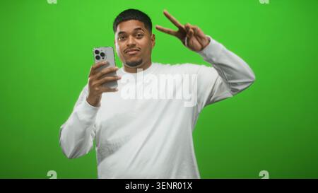 Mann hält Smartphone und zeigt Friedenszeichen mit der rechten Hand im grünen Studio, während er für Selfie lächelt; spielerische Inhaltserstellung. Stockfoto