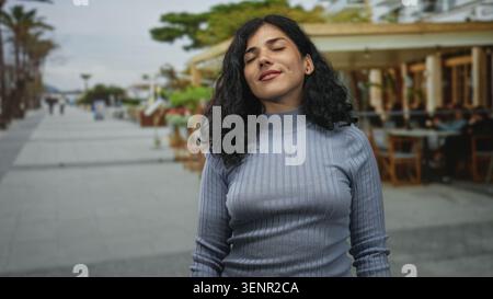 Junge brünette Frau, die mit geschlossenen Augen und einem leichten Lächeln auf der Straße neben einer Restaurantterrasse im Freien steht; ruhige Reflexion. Stockfoto