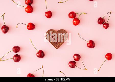 Frische Kirschen und herzförmige Schokolade auf hellrosa Hintergrund. Romantisches Essenskonzept. Flacher Style. Stockfoto