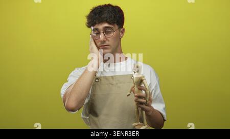 Junger Mann, der eine Holzpuppe hält und seine Wange in einem Atelier mit gelbgrünem Hintergrund berührt; Introspektion. Stockfoto