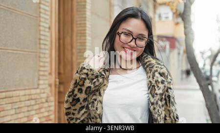 Frau mit Brille und Leopardenpelz lächelt mit der Hand hinter dem Kopf auf der Straße und lehnt sich an ein Gebäude; ungezwungenes Vertrauen. Stockfoto