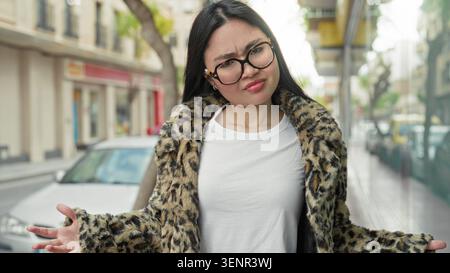 Frau im Leopardenmantel zuckt mit Handflächen auf der Straße, während sie den Kopf neigt, Brille und weißes T-Shirt trägt; Unklarheit. Stockfoto