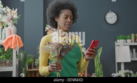 Floristin auf Schürze hält Blumen und Smartphone, lächelt beim Lesen des Telefons im Studio; kleine Geschäftsfreude. Stockfoto