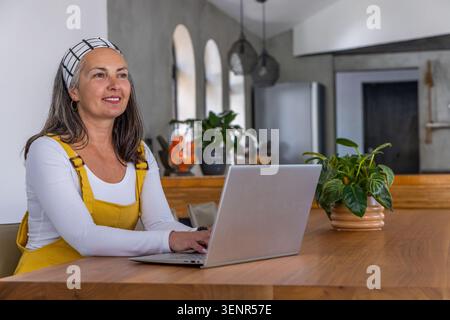 Senior-Frau, die an silbernem Laptop am Holztisch in der Küche arbeitet, Senfanzüge trägt, Pflanze Stockfoto