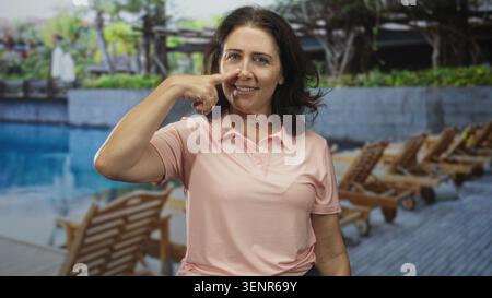 Eine Frau mit einem Lächeln zeigt Finger an Nase an den Liegestühlen am Pool und trägt im Studio eine pinkfarbene Polo-Platte; Entspannung und Wellness. Stockfoto