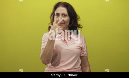 Frau zeigt mit dem Zeigefinger in gelbem Studio in rosafarbenem Poloshirt auf die Kamera; Anweisung: Ruhige Haltung. Stockfoto