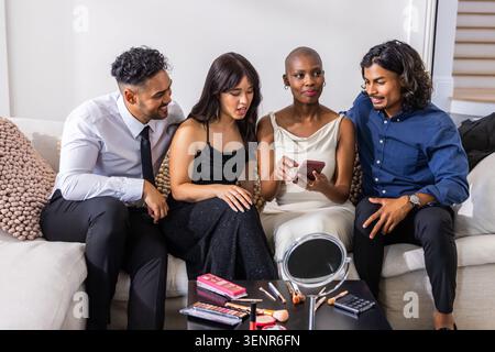 Verschiedene Freunde in formeller Kleidung sitzen im Wohnzimmer auf dem Sofa und teilen sich Telefon und Make-up-Paletten Stockfoto