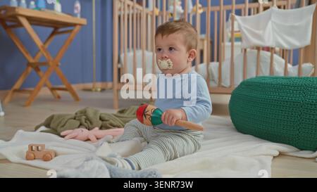 Junge, der auf einer Decke sitzt und eine hölzerne Maraca hält, Schnuller im Mund hält und ein Spielzeug mit der Hand hält, in einer Baumschule; sanfte Neugier. Stockfoto
