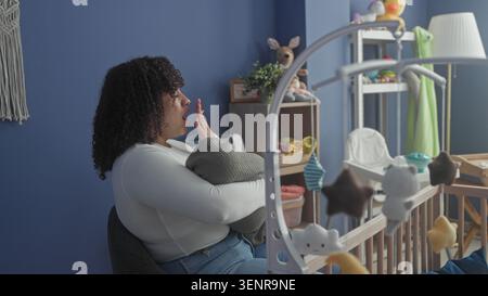 Die Frau wiegt das Baby in eine graue Decke, reibt die Augen und legt den Kopf neben dem Kinderbett mit einem gefüllten Häschen an die blaue Wand im Gebäude; Erschöpfungströst. Stockfoto