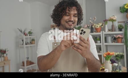 Mann Florist klopft Smartphone mit Händen in einem Blumenladen Gebäude, trägt eine Schürze und lächelt auf den Bildschirm; kleine Unternehmen kümmern sich gut. Stockfoto