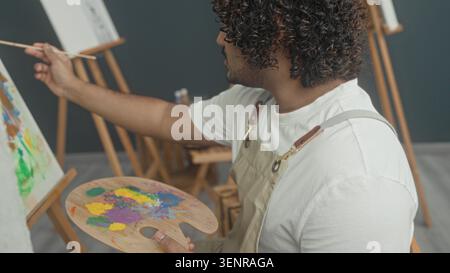 Junger indischer Mann in Schürze hält Farbpalette und Pinsel beim Malen auf Staffelei im Atelier, Farbflecken sichtbar auf Palette und Leinwand; ruhiger Konzentrat Stockfoto