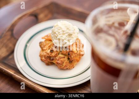 Ein gebackener Teig mit reichhaltiger Creme liegt auf einem Holztablett neben einem verschwommenen Eisgetränk und fängt einen gemütlichen entspannenden Café-Dessert-Moment perfekt ein Stockfoto