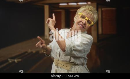 Frau in gelber Brille zeigt mit dem Finger nach oben und lächelt, während sie sich in einem dunklen Studio mit Holzbalken und -Dielen lehnt; Verspieltheit. Stockfoto