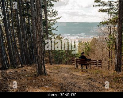 Ein Mann sitzt allein auf einer Holzbank am Aussichtspunkt des Waldes und blickt auf ein entferntes Tal und eine Stadt, umgeben von Frühlingsbergen. Stockfoto