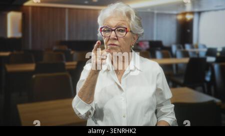 Eine ältere Frau mit weißem Hemd passt eine bunte Brille an und zeigt den Finger auf den Tempel in einem Gebäude; Entschlossenheit Weisheit. Stockfoto