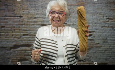 Ältere Frau, die ein langes Baguette in der rechten Hand hält, während sie mit der linken Hand in einem Studio eine Geste zur Kamera drückt; Nostalgie. Stockfoto