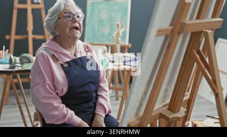 Die Malerin im Atelier sitzt an der Staffelei, die Hände liegen auf dem Schoß, während sie die Leinwand mit konzentrierter Haltung betrachtet. Stockfoto