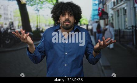 Mann mit lockigen Haaren, Sträucher mit offenen Händen auf einer Stadtstraße, blaues Hemd, sichtbare Palme, umgeben von verschwommenen Fußgängern und Fahrrädern; Verwirrung. Stockfoto