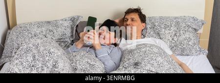 Ein verheiratetes Paar wacht an einem typischen Morgen im Bett auf. Die Frau benutzt ein Telefon, während der Mann neben ihr liegt. Sie teilen einen gemütlichen Moment in ihrem Be Stockfoto